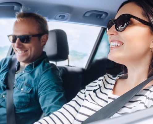 A happy woman driving in a car with a male passenger looking at his phone.